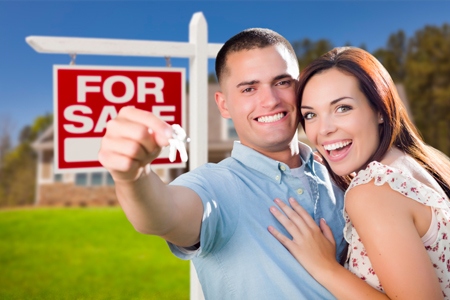 Mixed Race Excited Military Couple In Front of New Home with New House Keys and For Sale Real Estate Sign Outside.